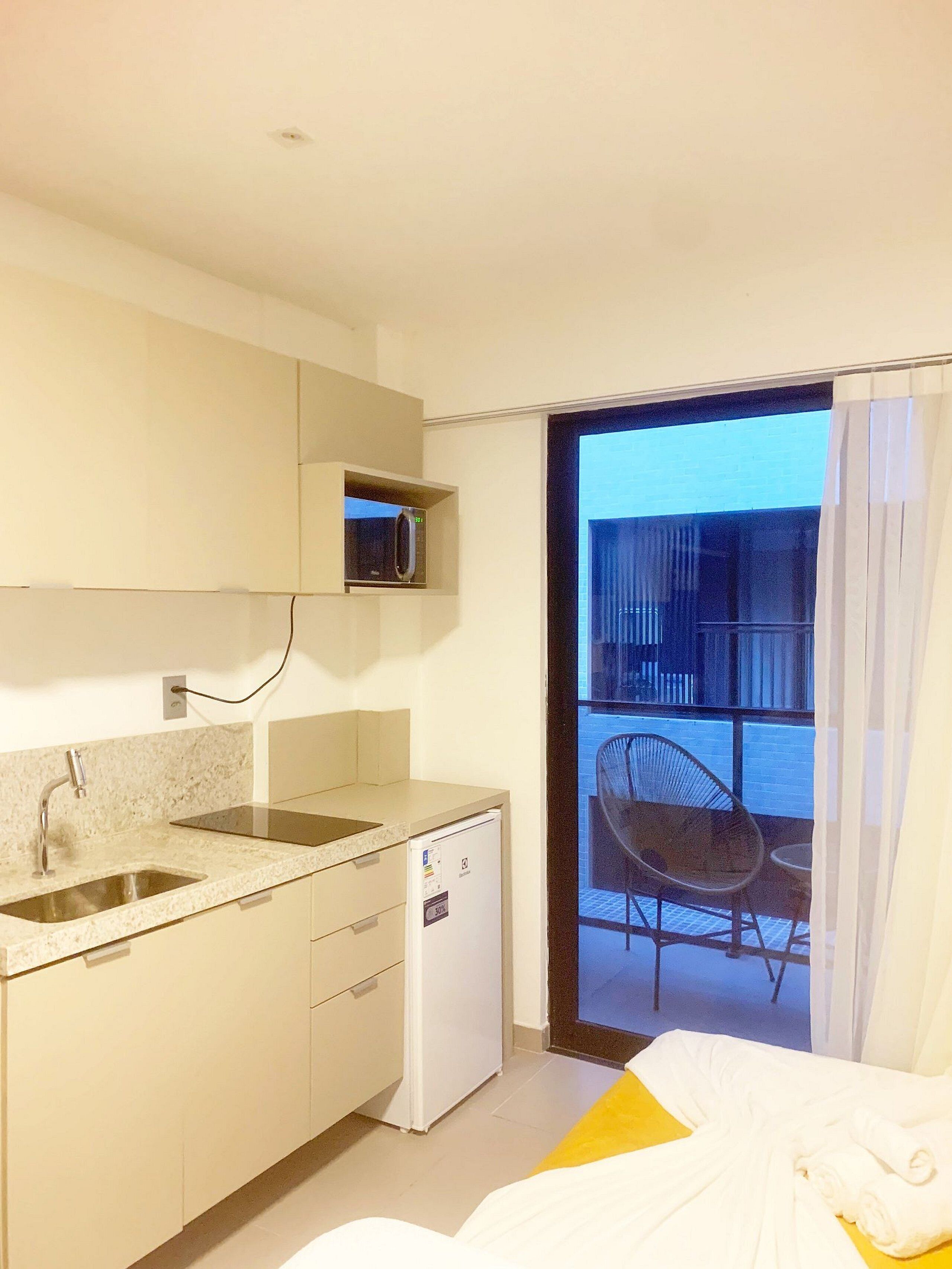 Een compact keukenhoekje met een magnetron, een kleine koelkast en een spoelbak. Er is een balkon met een stoel en een tafel, en een bed met wit beddengoed.