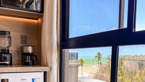 A kitchen with a refrigerator, coffee maker, and blender. A dining table with a chair is in front of a window with a view of buildings and palm trees. - Beach Haus by Day by Day (Joao Pessoa)