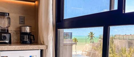 A kitchen with a refrigerator, coffee maker, and blender. A dining table with a chair is in front of a window with a view of buildings and palm trees.