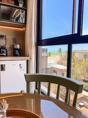 Una cocina con refrigerador, cafetera y licuadora. Un comedor con una silla está frente a una ventana con vista a edificios y palmeras.