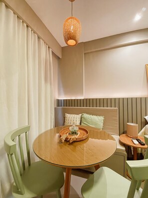 A modern dining area with a round wooden table, green chairs, a woven pendant light, and a bed with a headboard and pillows in the background. - Beach Haus by Day by Day (Joao Pessoa)
