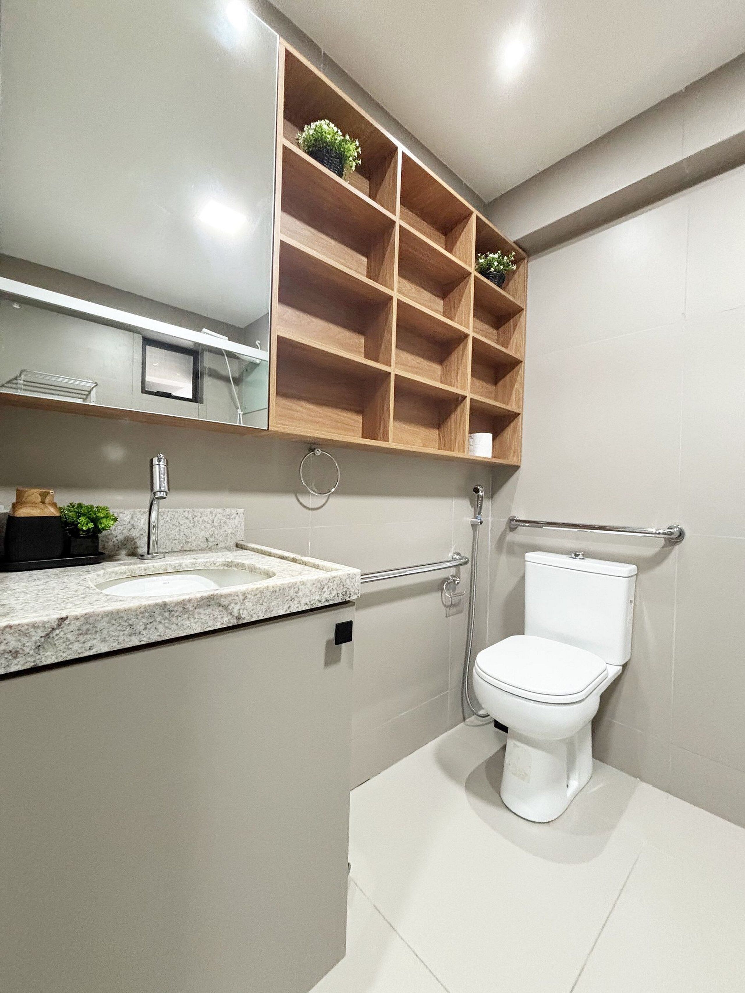 Um banheiro moderno com bancada de granito, vaso sanitário, prateleira de madeira com plantas e espelho sobre a pia.