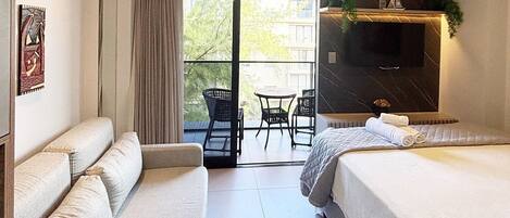 Hotelkamer met een bank, een bed, een televisie, een balkon met stoelen en uitzicht op groen.