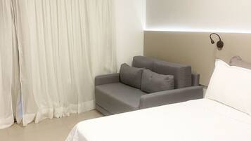 Habitación de hotel con cama, sofá, mesita de noche y ventana con cortinas traslúcidas.