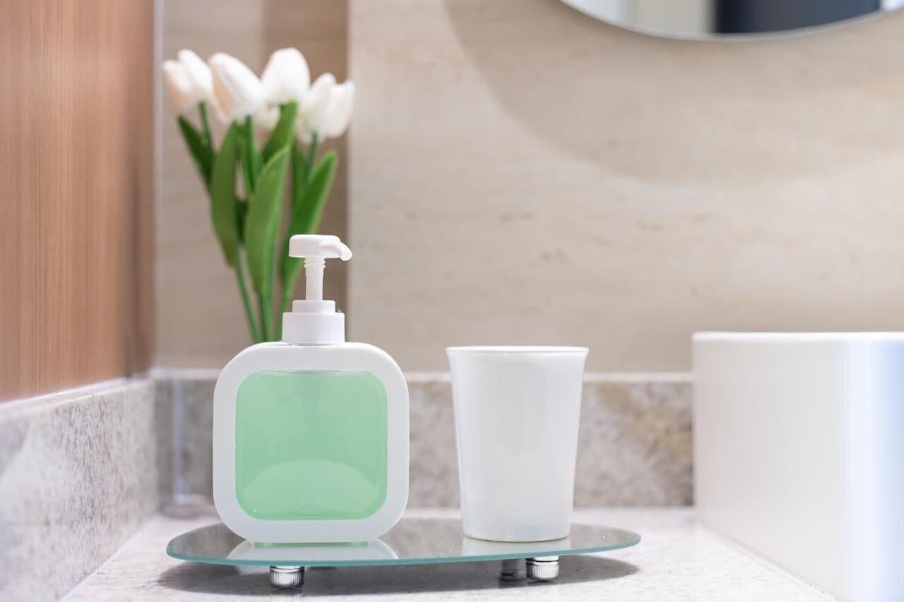 Un baño con dispensador de jabón de vidrio, una taza blanca y un florero con flores blancas.