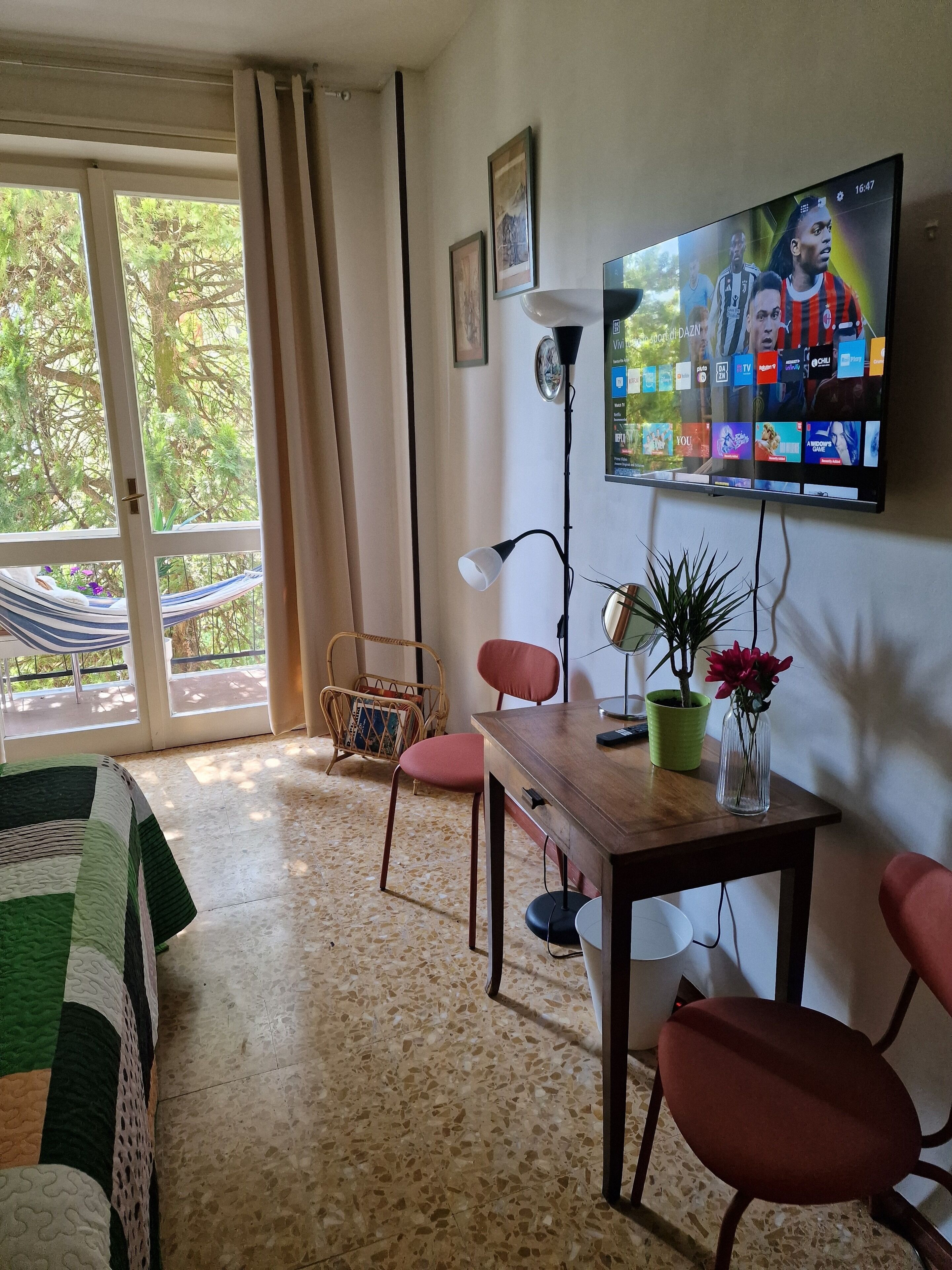 A living room with a TV displaying a sports channel, a dining table with chairs, a floor lamp, and a view of a balcony with a hammock.