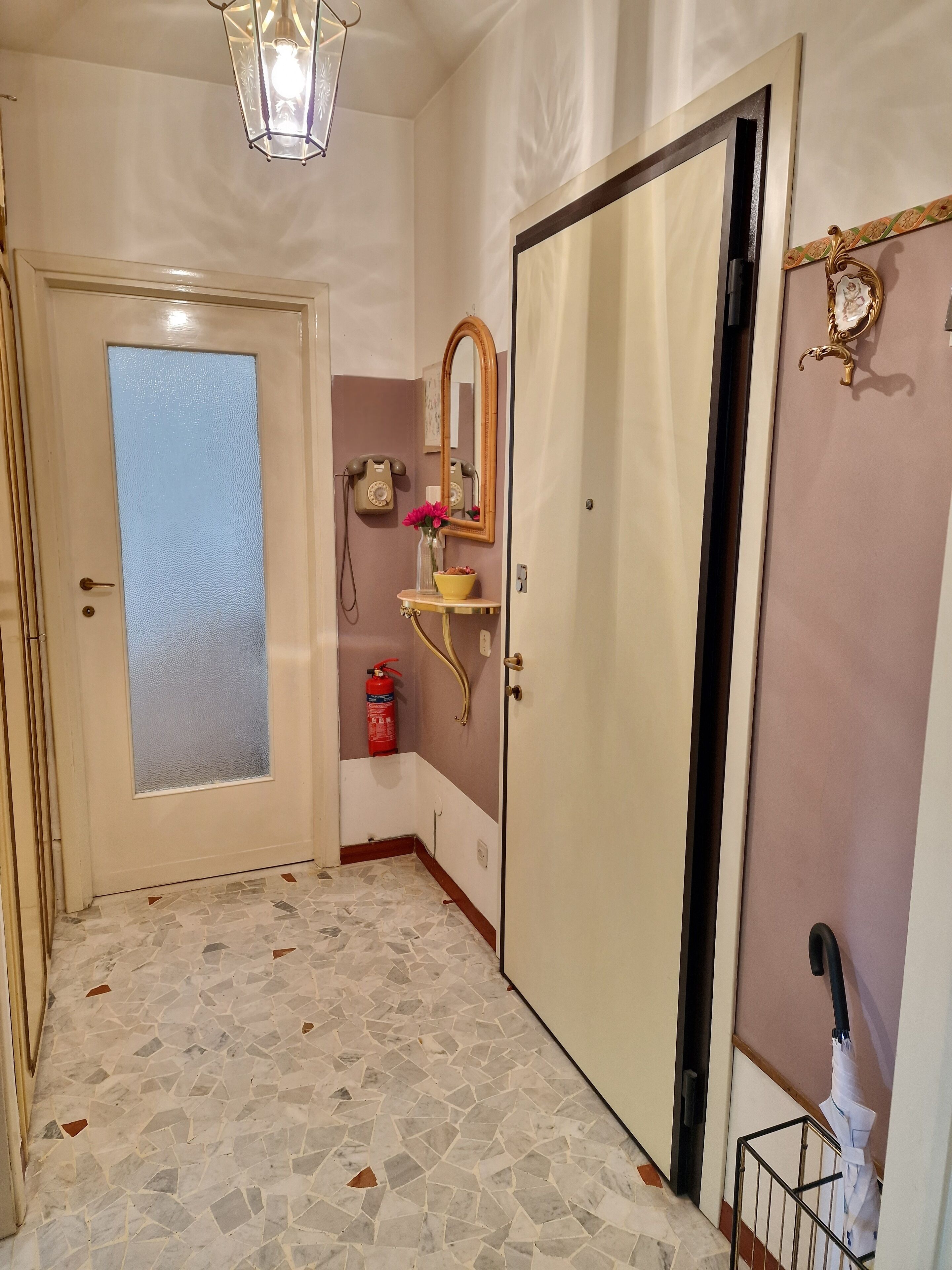 A hallway with a tiled floor, a door with frosted glass, a wall-mounted telephone, a mirror, a small table with a vase, and a fire extinguisher.