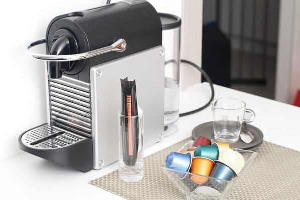 A coffee machine with a drip tray, a glass of water, and a container with colorful cups.
