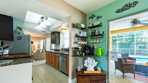 A kitchen with a coffee bar, a dining area with a pool view, and a living room with a sofa and curtains.
