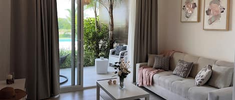 A modern living room with a sofa, a coffee table, and a view of a pool and greenery through large windows.