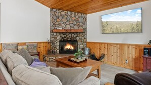 A cozy living room with a stone fireplace, wooden ceiling, and a large window overlooking a scenic landscape.