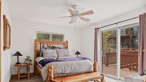 A bedroom with a wooden bed, a ceiling fan, a wooden bench, a nightstand with a lamp, and a large window with a view of a deck.