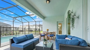 A covered patio with wicker furniture, a glass table, and a view of a swimming pool and houses.