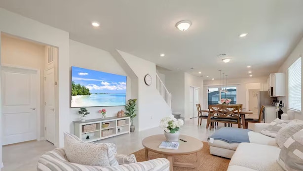 A spacious living area with a large flat-screen TV mounted on the wall, a dining area with a wooden table and chairs, and a kitchen area with white cabinets.