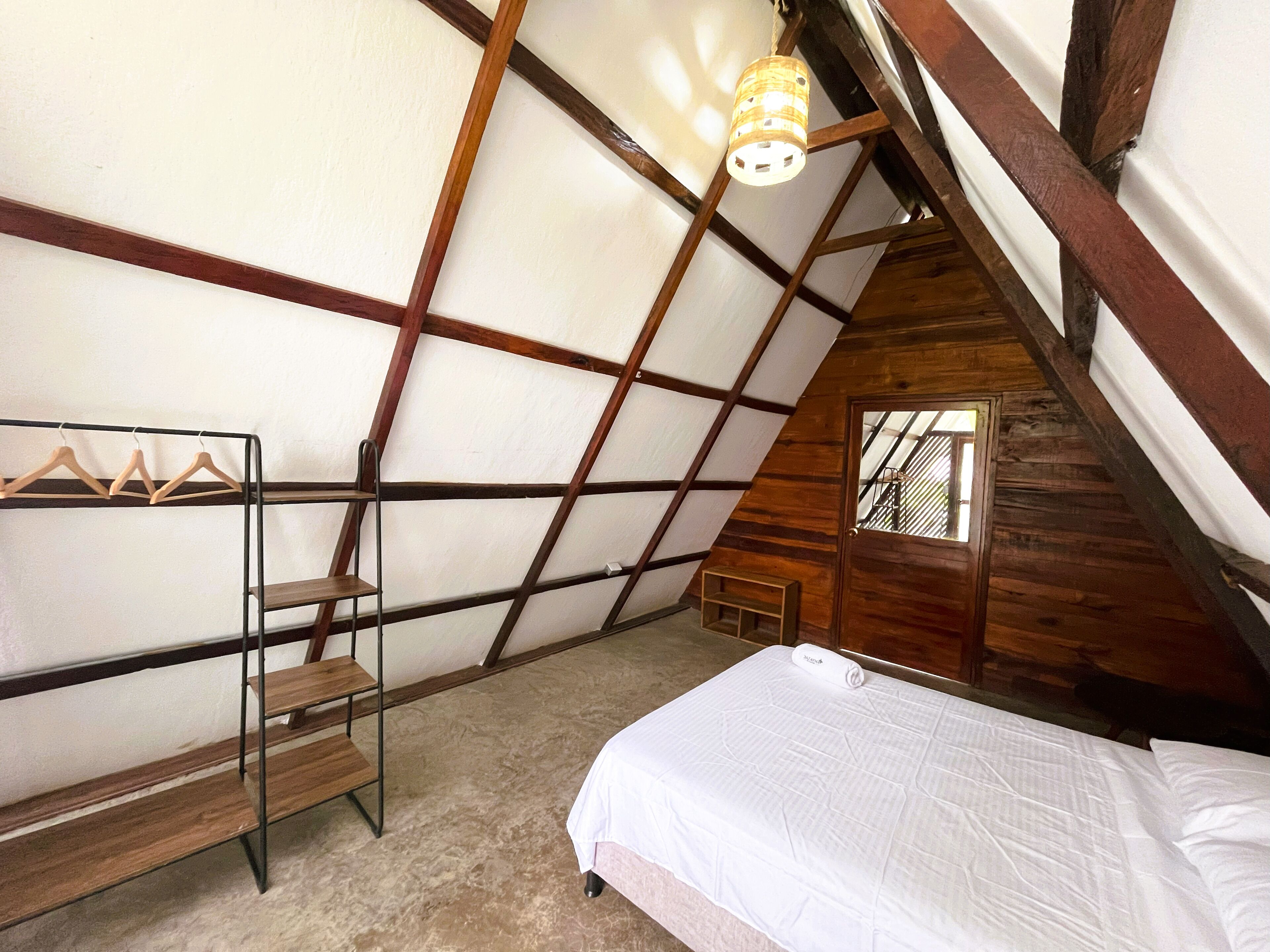 Une chambre avec un plafond en pente, des poutres en bois, un lit, un luminaire suspendu et une fenêtre donnant sur un beau paysage.