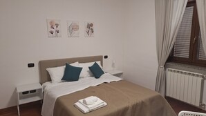 A neatly made bed with white and teal pillows, a brown blanket, and a folded white towel on the bed. There are two bedside tables with lamps, a wall-mounted control panel, and three framed artworks above the bed.