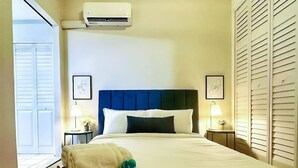 Ein Hotelzimmer mit einem großen Bett, zwei Nachttischen mit Lampen, einer wandmontierten Klimaanlage und Blick ins Freie durch eine Tür mit Fensterläden.