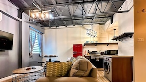 A modern living room with a sofa, a round coffee table, and a TV mounted on the wall. A kitchen area with a washing machine and a dining table is visible in the background.