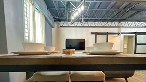 A modern dining area with a long table, four cushioned chairs, a flat-screen TV, and a ceiling with exposed beams and lighting fixtures. - Lofts @ Santurce – Your Urban Hideaway (San Juan)