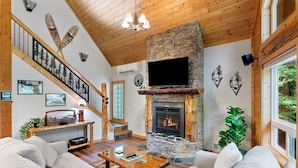 Una sala de estar acogedora con chimenea de piedra, techo de madera y una escalera con barandilla decorada con motivos forestales.