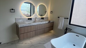 Un baño moderno con una bañera independiente, un lavabo doble con espejos redondos y un ventanal que proporciona luz natural.