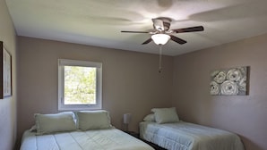 Un dormitorio con dos camas, un ventilador de techo, una ventana con vistas a árboles y un cuadro colgado en la pared.