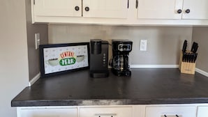 Una cocina con armarios blancos, una cafetera, un cartel enmarcado que dice 'Central Perk' y un juego de cuchillos.