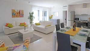 A modern living room with white sofas, a dining table set for two, and a kitchen in the background. - Gorgeous home in Rebici (Rebici)