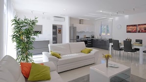 A modern living room with a white sofa, a coffee table, and a dining area with a table and chairs. There is a kitchen in the background with cabinets and a refrigerator. - Gorgeous home in Rebici (Rebici)
