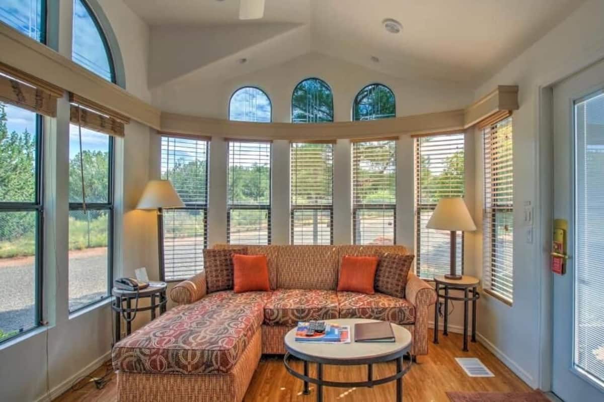 A spacious living room with a large sectional sofa, a round coffee table, and two lamps. Large windows provide ample natural light and views of the outdoors.
