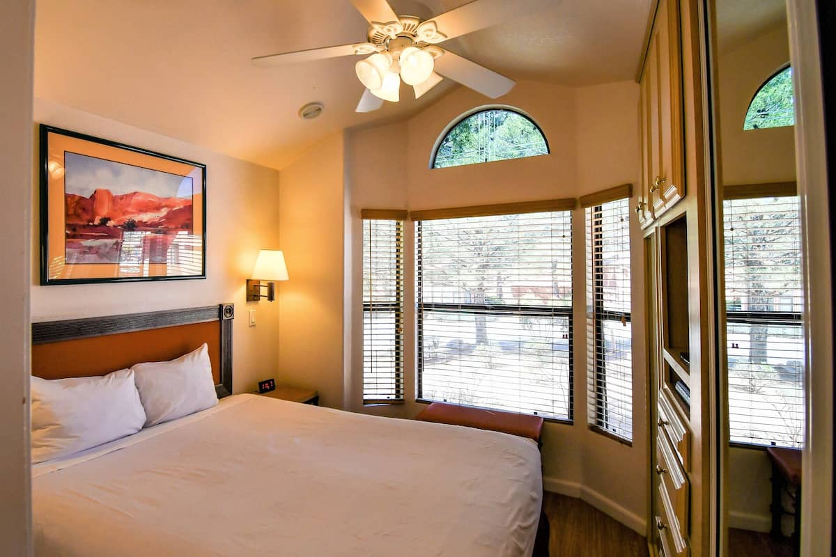 A hotel room with a bed, a ceiling fan, a painting, a lamp, and large windows with blinds.