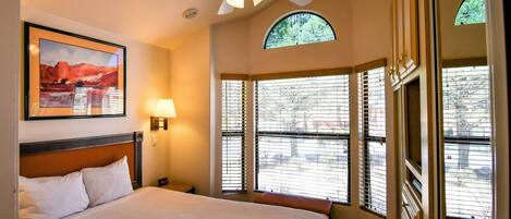 A hotel room with a bed, a ceiling fan, a painting, a lamp, and large windows with blinds.