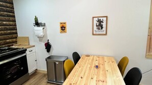Une table de salle à manger en bois, accompagnée de quatre chaises, une poubelle, une petite étagère avec une bouteille et un tableau encadré accroché au mur.