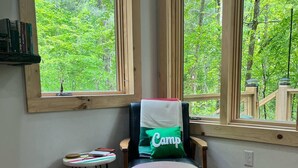 Une pièce dotée de grandes fenêtres, d’un plancher en bois, d’une chaise avec un coussin orné du mot « Camp », d’une petite table et d’une bibliothèque.