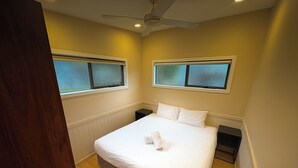 Un dormitorio con una cama, dos mesitas de noche, un ventilador de techo y ventanas con vidrio esmerilado.