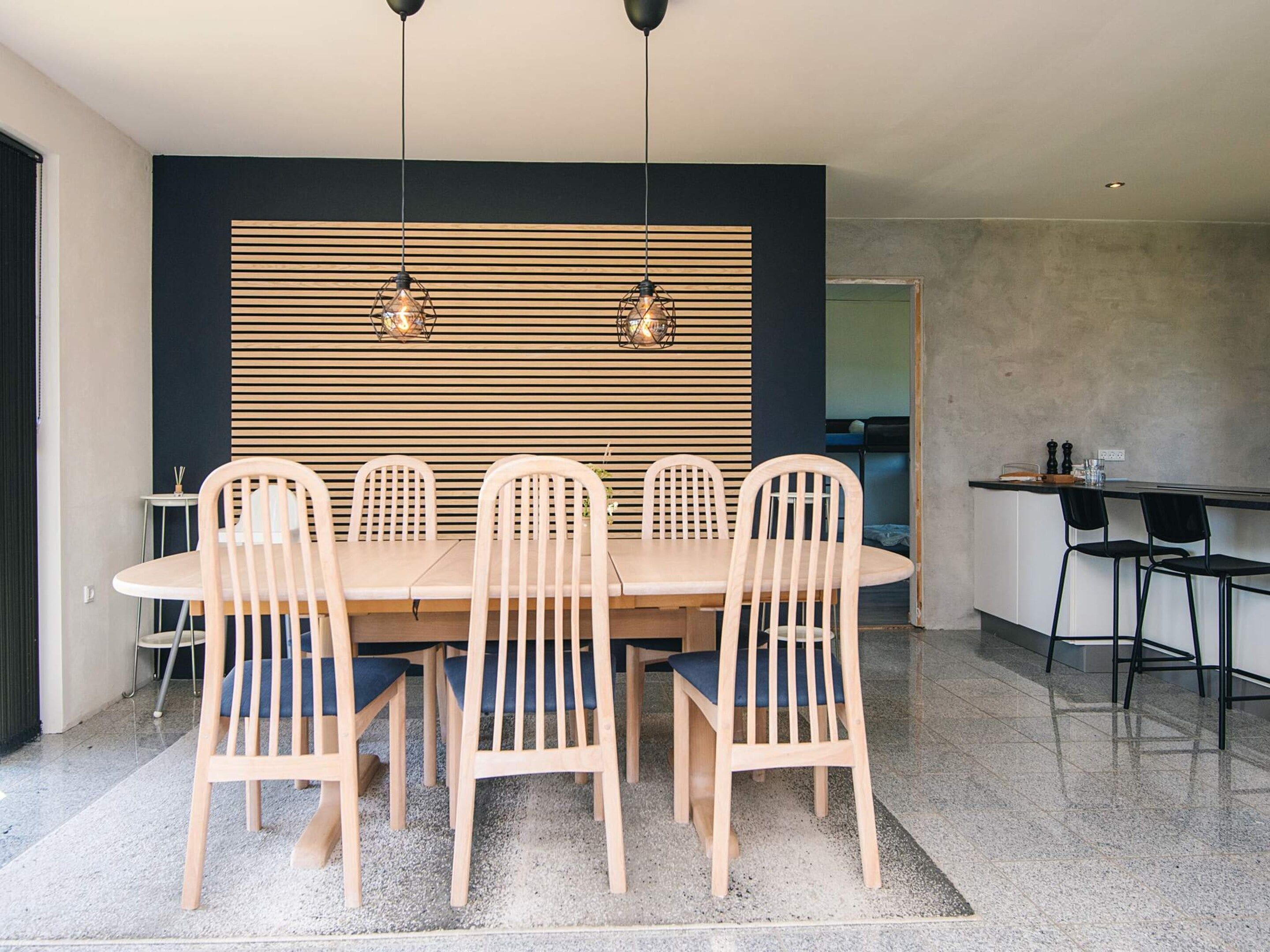 A dining area with a wooden table and chairs, a black wall with horizontal slats, and pendant lighting.