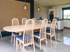 A modern dining area with a round table and six chairs, a kitchen in the background, and pendant lighting.