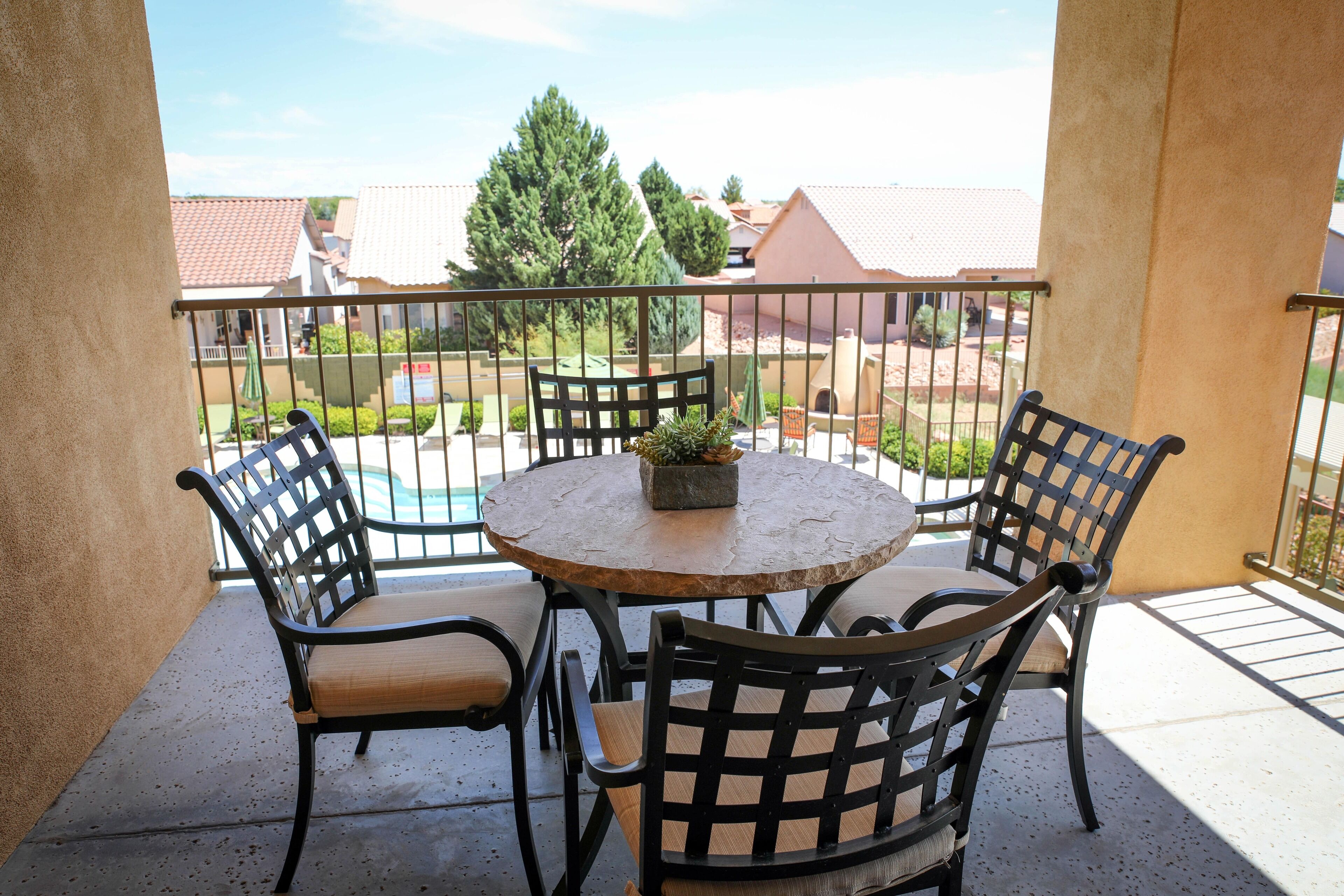 A balcony with a round table and four chairs, a potted plant, and a view of a swimming pool and buildings.