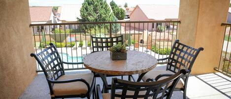 A balcony with a round table and four chairs, a potted plant, and a view of a swimming pool and buildings.