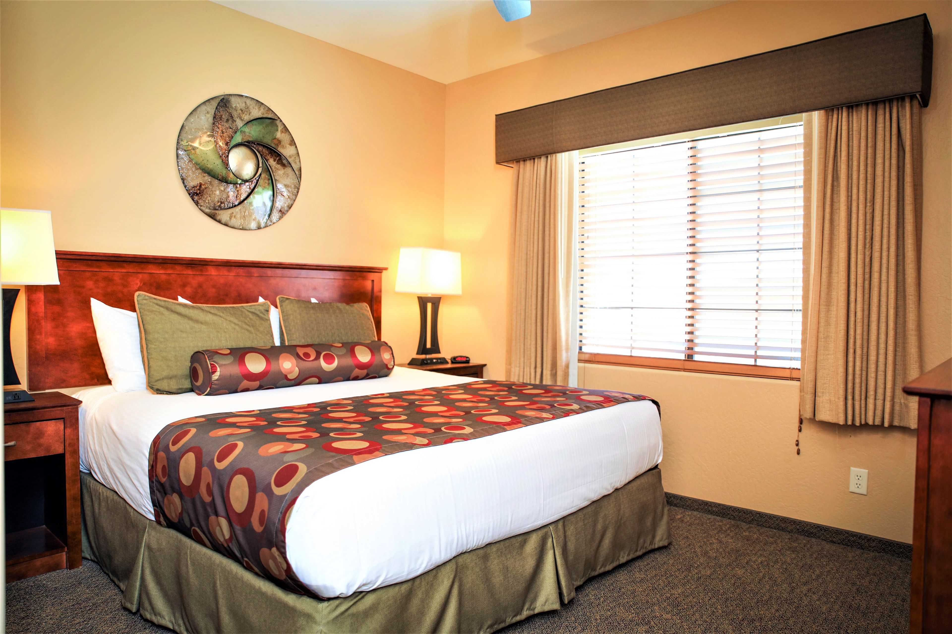 A hotel room with a large bed, two bedside tables, a wall clock, a window with blinds, and a decorative wall piece.