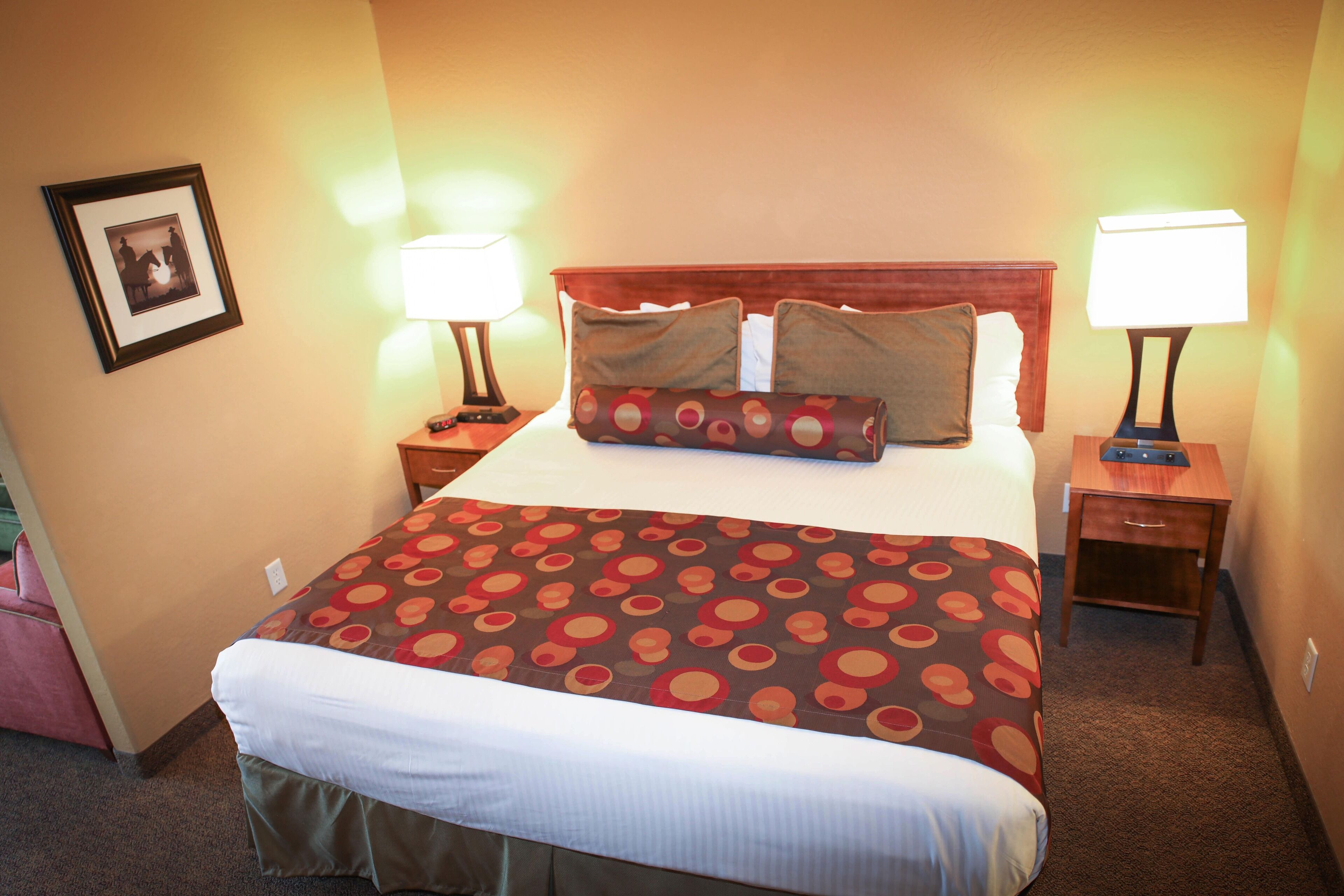 A hotel room with a large bed, two bedside tables with lamps, a framed picture on the wall, and a carpeted floor.