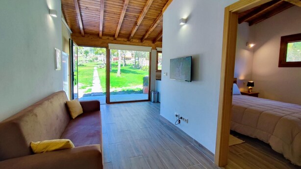 Ein Zimmer mit Holzboden, einer Couch, einem Bett, einem Fernseher und Blick auf einen Garten durch eine Schiebetür.