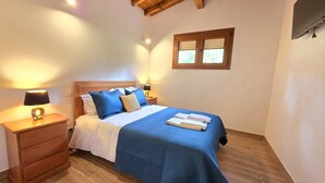 Ein Schlafzimmer mit Holzboden, einem Bett mit blauer Bettwäsche, einem Nachttisch mit Lampe und einem Fenster mit Blick auf Grünflächen.