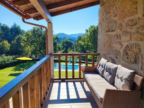 A balcony with a stone wall, a wooden railing, a view of a pool, and a grassy area with trees.
