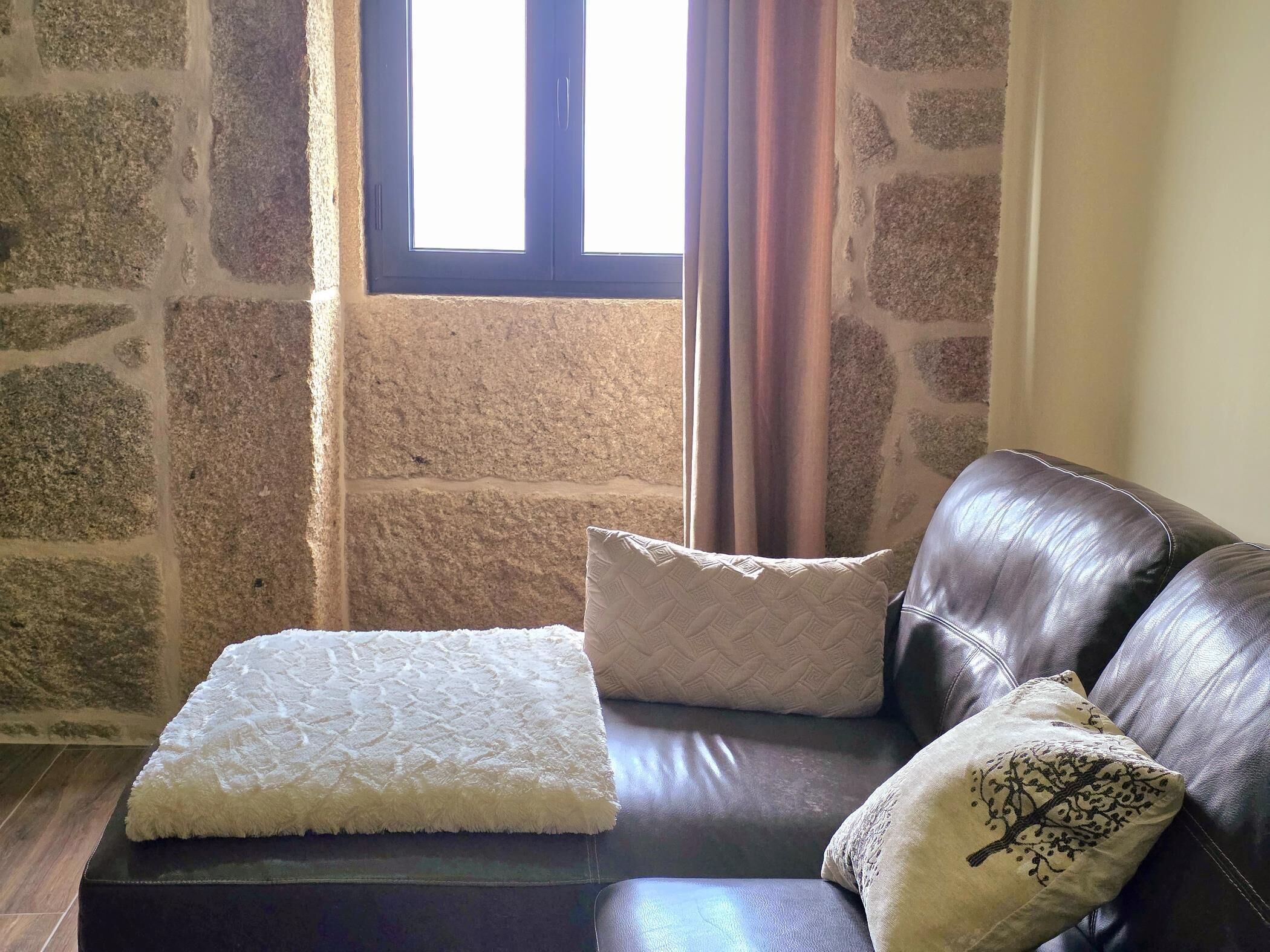 Una habitación con pared de piedra, ventana con cortinas, sofá de cuero con cojines y una alfombra blanca y mullida.