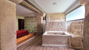 A stone-walled room with a wooden bench and a red table, a stone fireplace, and a window.