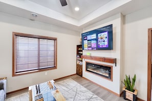 Una sala de estar con un televisor de pantalla plana montado en la pared, chimenea, una ventana grande con persianas y una mesa de centro.