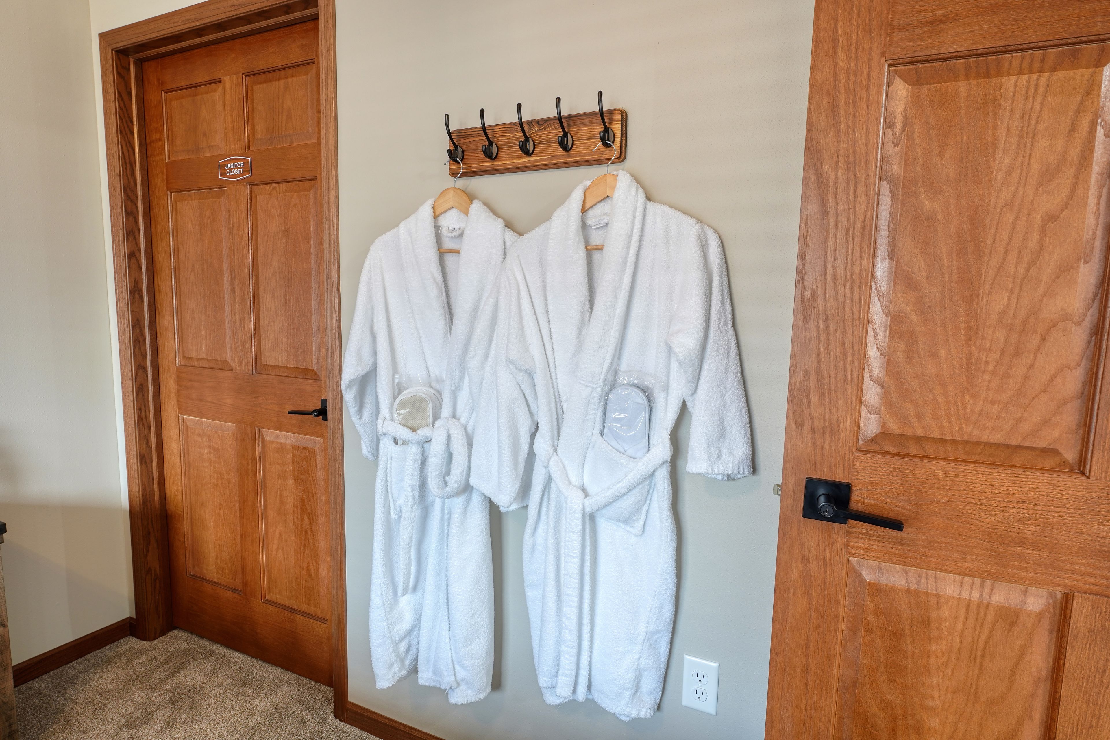 Two white bathrobes hanging on a wooden rack in a hallway with wooden doors.