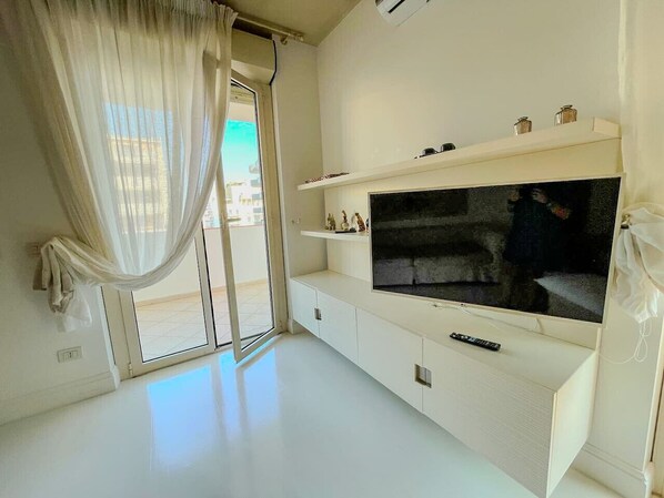 Ein modernes Wohnzimmer mit großem Flachbildfernseher, einer Schiebetür zum Balkon und einer weißen Unterhaltungsanlage.