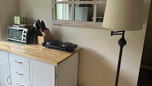 A kitchenette with a microwave, toaster, and knife block on a wooden countertop, a white cabinet with drawers, a floor lamp, and a window with a mirror reflecting a coffee maker and a refrigerator. - Piermont Oasis (Piermont)